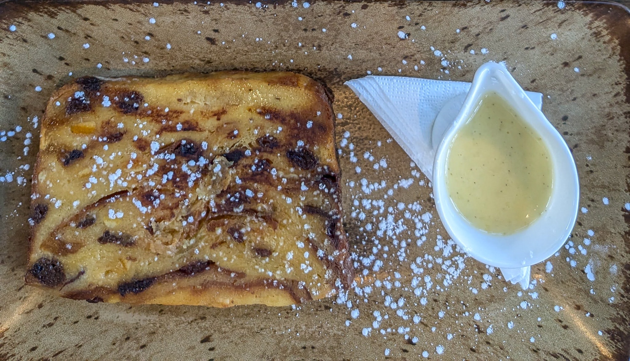 A flatlay of a slice of panettone bread and butter pudding sprinkled with icing sugar, and a small white jug of whiskey custard.