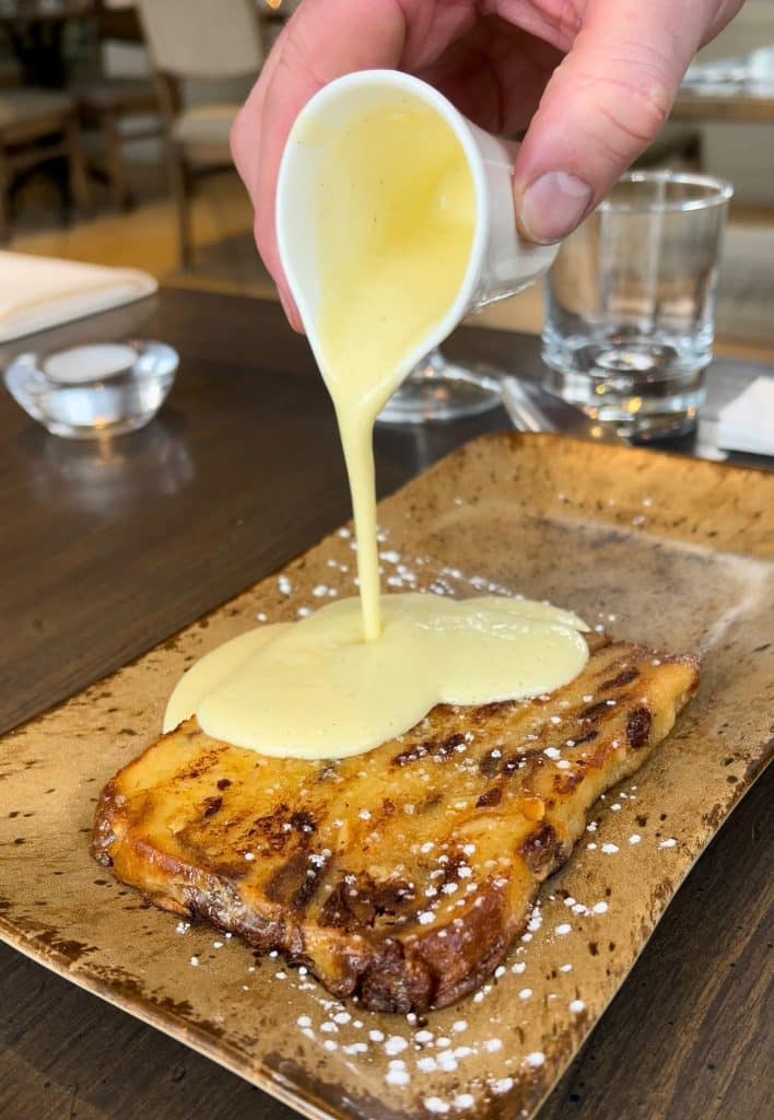 A small white jug of whiskey custard is poured over a slice of panettone bread and butter pudding.