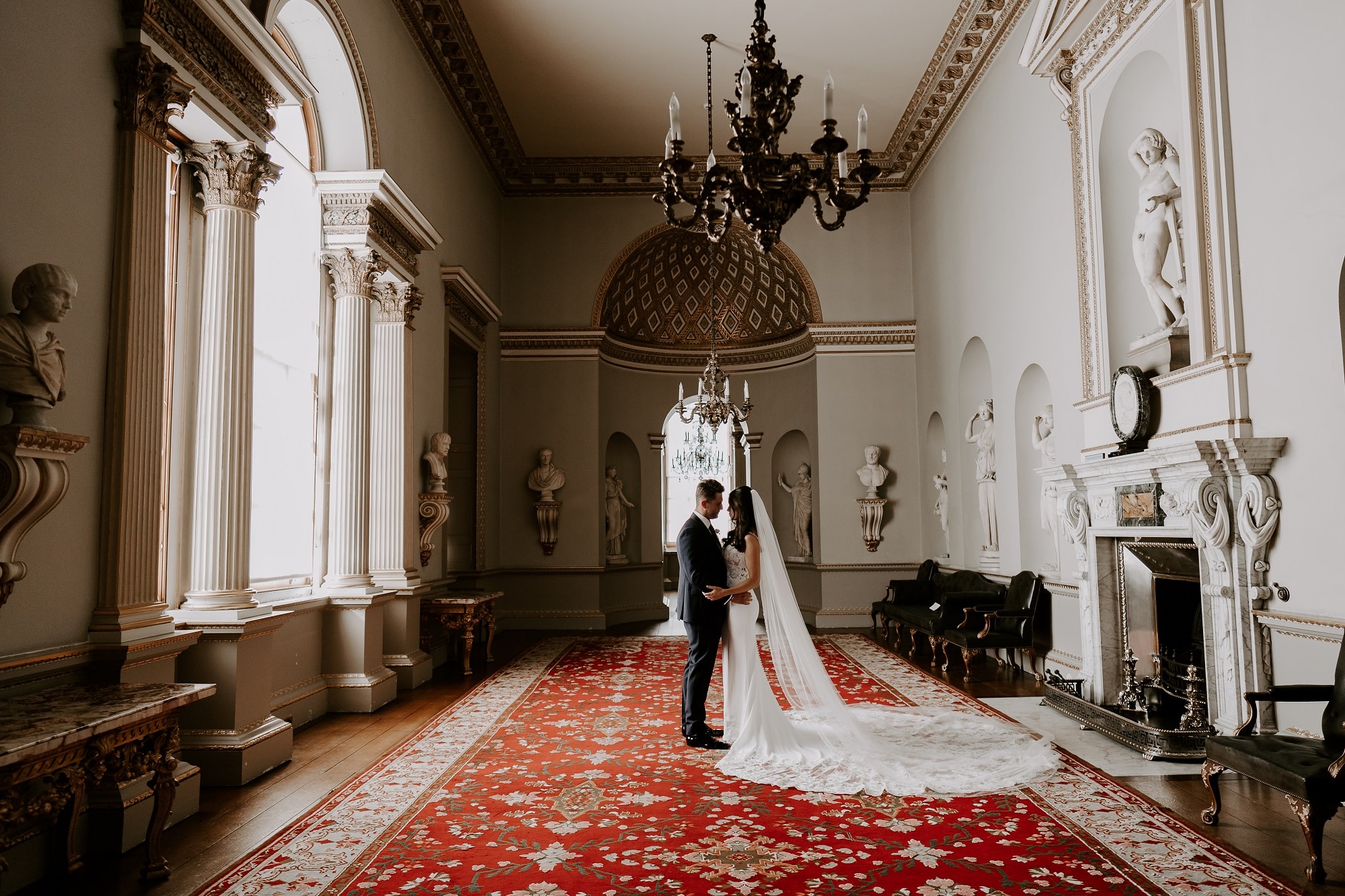 A bride and groom gaze into each other's eyes in the Statue Gallery of Holkham Hall, an impressive wedding venue and stately home in Norfolk. A patterned, red carpet lies on the floor while Roman and Greek statues stand in arches on the walls.