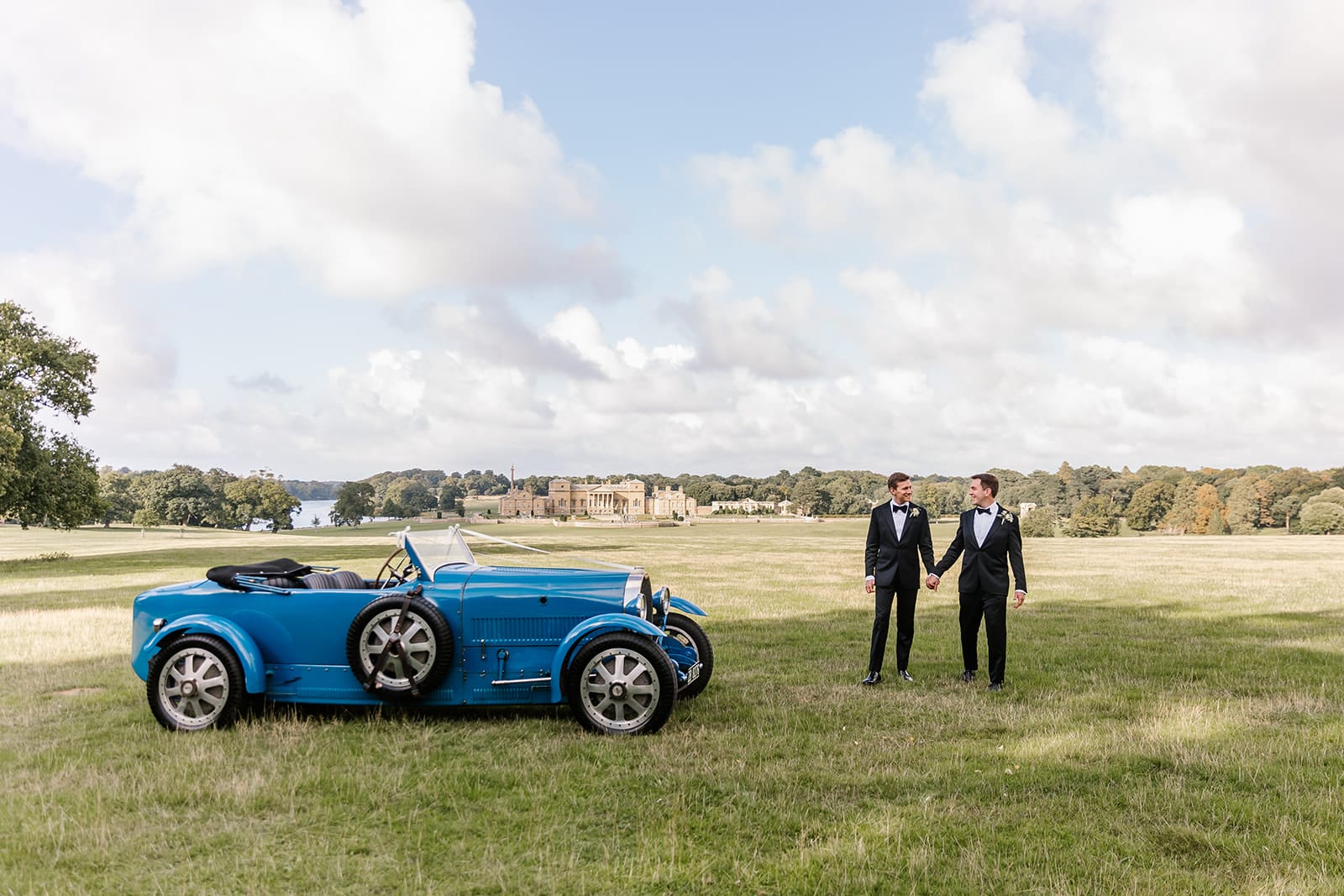 Two grooms stand holding hands next to a blue vintage open top car, with Holkham Hall in the background.