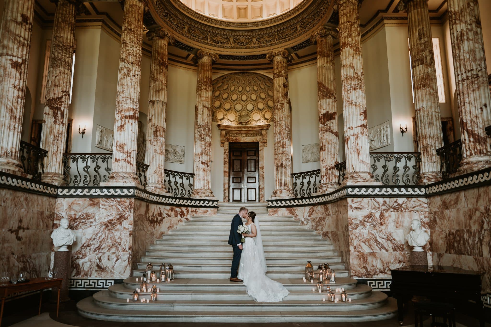 A bride and groom kiss as they stand on the steps of the Marble Hall at Holkham, a stately home wedding venue.