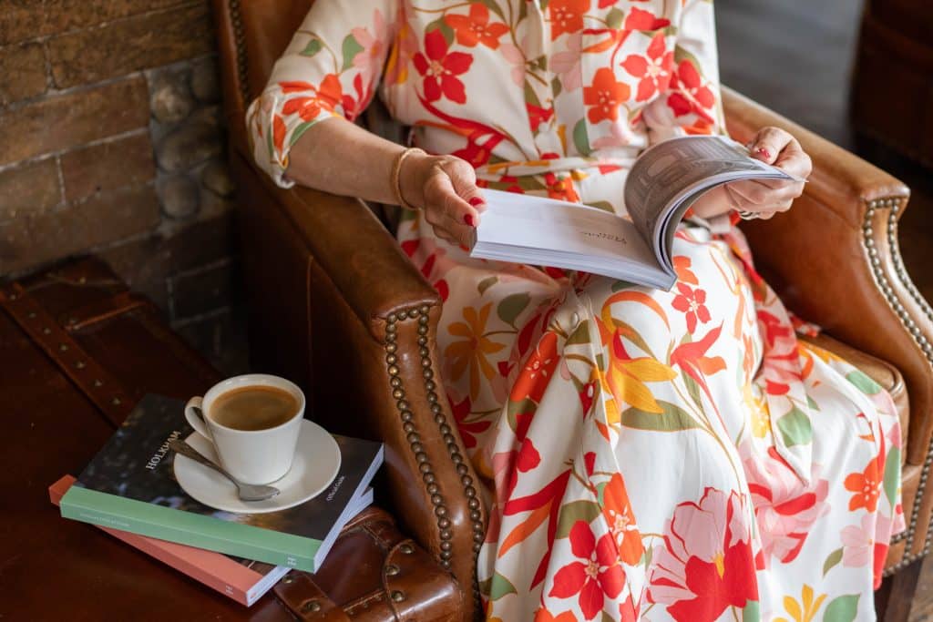 A lady in a colourful floral dress reads a book, sitting in a leather armchair. A cup of coffee stands on a stack of books beside her.