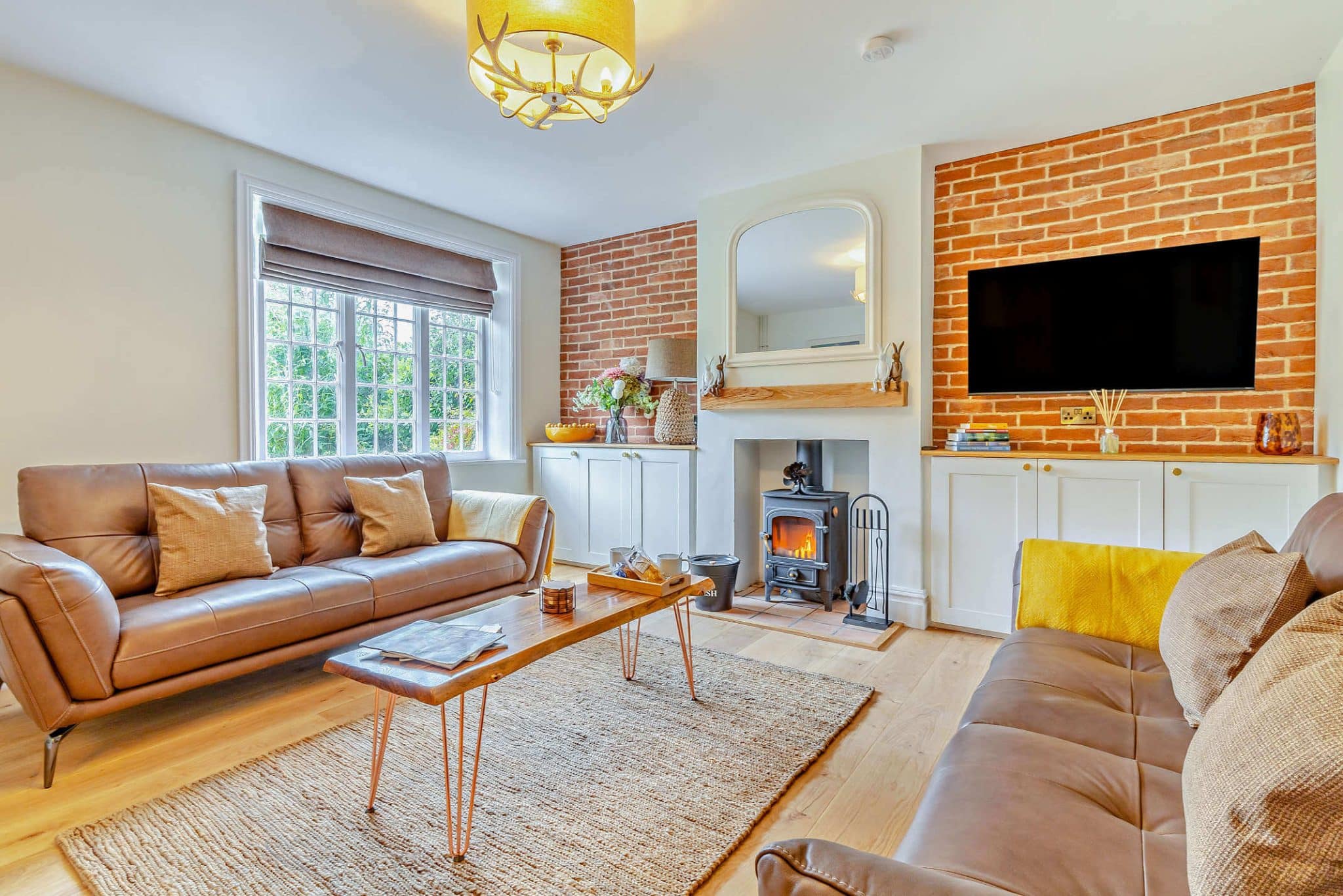 A bright, modern holiday home lounge area with leather sofas, a wood burner and a TV.