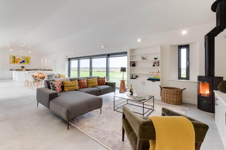 A large open plan living area with grey L shape sofa, wood burner, and kitchen in the background.